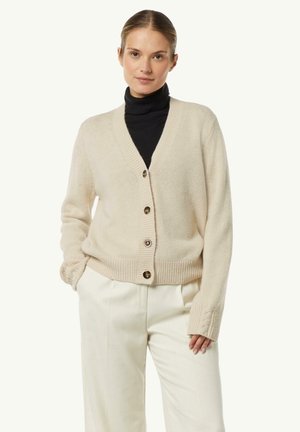 Beige cardigan with a V-neck, five brown button closures, ribbed cuffs, and hem. Worn over a black turtleneck. Soft knit texture.