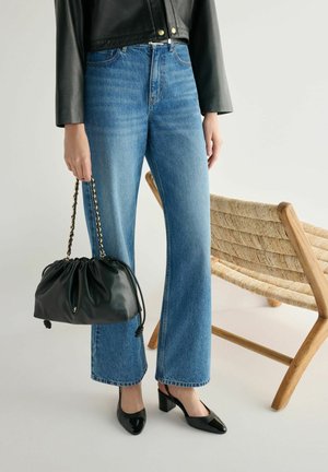 Osoba ubrana w niebieskie dżinsy i czarną skórzaną kurtkę, trzymająca czarną torebkę na łańcuszkowym pasku, stojąca obok plecionego drewnianego krzesła.