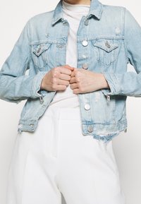 Veste en denim bleu clair avec des détails usés, deux poches poitrine, boutons en métal argenté et longueur cropped.