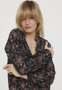 Blouse à motifs floraux en violet foncé et noir. Dotée d'un tissu texturé, d'une fermeture à boutons et d'un col à volants. Manches longues avec des poignets élastiques.