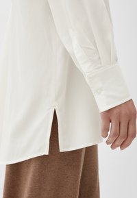 Biała koszula z długim rękawem, z bocznym rozcięciem na dole oraz zapięciem na guzik przy mankietach. Gładki materiał o lekko usztywnionej strukturze.