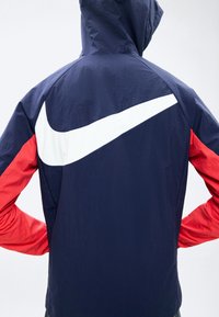 Άτομο που φοράει σκούρο μπλε κουκούλα με κόκκινα μανίκια και μεγάλο λευκό λογότυπο Nike Swoosh στην πλάτη.