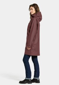 Veste imperméable bordeaux avec capuche, poches latérales et manches longues. Portée sur un jean bleu et des bottines noires, présentant une texture lisse.