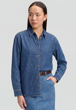 Frau trägt ein dunkelblaues Jeanshemd mit Knöpfen und einen hellen Jeansrock mit braunem Gürtel, steht mit einer Hand in der Tasche.