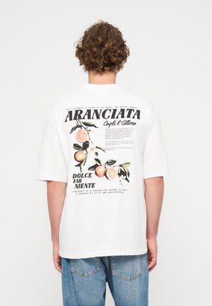 Homme aux cheveux bouclés portant un t-shirt blanc avec une illustration de fruits d'agrumes et un texte italien au dos, associé à un jean bleu.