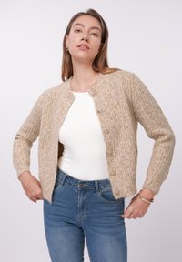 Cardigan beige de punto con textura acanalada, escote redondeado y seis botones transparentes, llevado sobre una camiseta blanca y combinado con vaqueros azules.