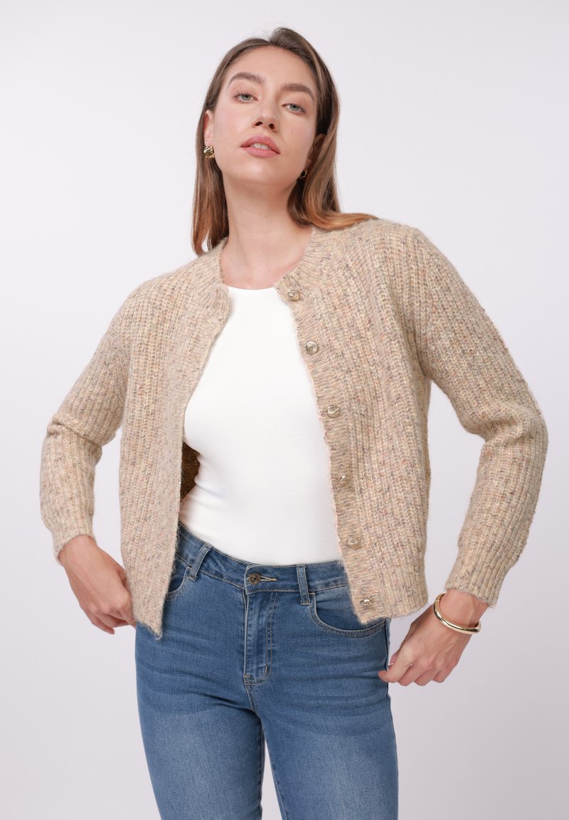Cardigan beige de punto con textura acanalada, escote redondeado y seis botones transparentes, llevado sobre una camiseta blanca y combinado con vaqueros azules.