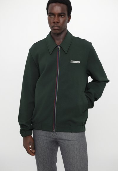 Mørkegrønn zip-jakke med bred krage, rød- og blåstripet glidelås og en liten logo-patch på venstre bryst. Myk stoff.