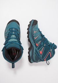 Tyrkysové turistické boty s černými akcenty, vybavené texturovaným svrchním materiálem, síťovanými panely a robustními gumovými podrážkami. Jasně oranžové detaily podél stran.