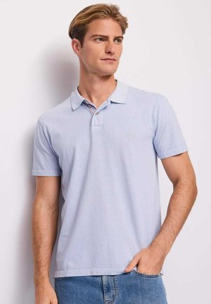 Giovane uomo con capelli chiari che indossa una polo azzurro chiaro a maniche corte e jeans, in piedi con una mano in tasca, guarda verso destra.