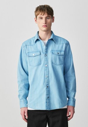 Jauns vīrietis ar īsiem brūniem matiem, valkājot gaiši zilu džinsa krekliņu ar pogām virs baltas topiņas un melnām biksēm, pret vienkāršu fona.