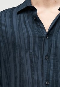 Skjorte med krage i mørkeblått stoff med vertikale, teksturerte striper og diskret glans. Har fem knapper foran og en ledig passform.