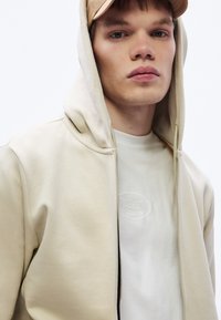 Beżowa bluza z kapturem na zamek błyskawiczny z sznurkiem w kapturze, noszona na białej koszuli z wytłoczonym logo. Gładka tekstura z luźnym krojem.