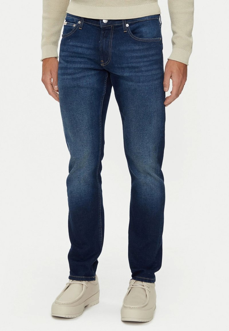 Jeans de mezclilla azul oscuro con un corte ajustado, que cuentan con un clásico diseño de cinco bolsillos, costuras visibles y un sutil efecto de desgaste.