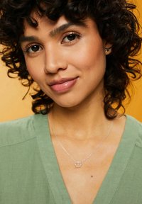 Collana d'argento con un pendente circolare adornato da piccoli accenti rotondi, indossata sopra un top di tessuto verde su uno sfondo arancione.