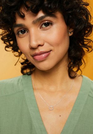 Collana d'argento con un pendente circolare adornato da piccoli accenti rotondi, indossata sopra un top di tessuto verde su uno sfondo arancione.