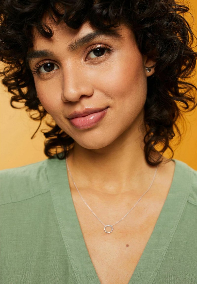 Collana d'argento con un pendente circolare adornato da piccoli accenti rotondi, indossata sopra un top di tessuto verde su uno sfondo arancione.