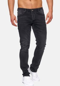 Musta farkkukangas, jossa on slim fit -määritys, viisi taskua, hienovarainen kulutusjälki ja metallinappikiinnitys, yhdistetty valkoisiin urheilukenkihin.