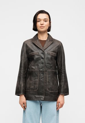 Mladý človek s krátkymi tmavými vlasmi, oblečený v zapnutú opotrebovanú čiernu koženú bundu a svetlomodré nohavice, stojaci pred jednoduchým bielym pozadím.