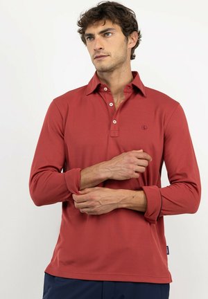 Hombre con cabello corto y rizado que lleva una camisa polo de manga larga color rojo óxido, ajustándose la manga y mirando ligeramente hacia un lado sobre un fondo blanco liso.