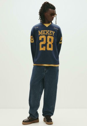 Navy long-sleeve jersey with yellow "MICKEY" and number "28" graphics, paired with loose-fitting blue denim jeans and brown shoes.