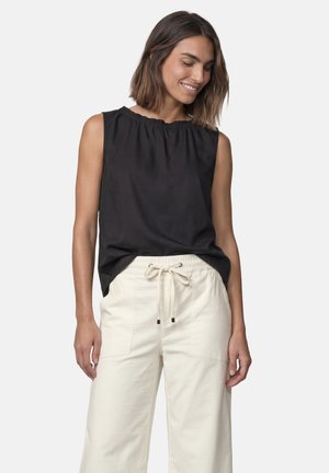 Femme aux cheveux mi-longs portant un haut noir sans manches et un pantalon crème à coulisse, souriante et regardant vers le bas sur le côté.