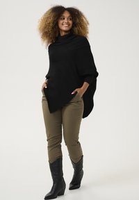Poncho nero a maglia con linee testurizzate, abbinato a pantaloni khaki e stivali alla caviglia neri, caratterizzato da un collo alto attillato e una silhouette rilassata.