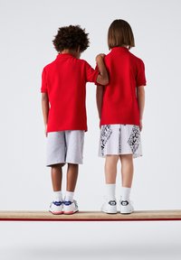 Due bambini sono in piedi su una tavola di legno, indossando camicie rosse con colletto. Un bambino ha pantaloni corti grigi, l'altro indossa pantaloni corti bianchi a fantasia. Entrambi indossano scarpe da ginnastica.