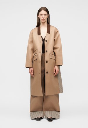 Femme portant un long manteau beige avec un col marron foncé, un pantalon à jambes larges avec des revers à carreaux, et des chaussures noires, debout devant un fond uni.