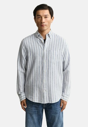 Uomo con camicia a righe verticali bianche e blu abbottonata sopra una maglietta nera, abbinata a jeans blu, in piedi davanti a uno sfondo uniforme.