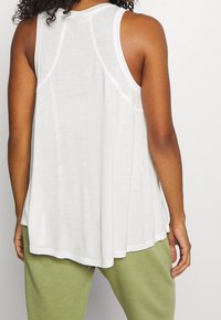 Bílý bezrukávový top vyrobený z lehké tkaniny, s volným designem a zaobleným lemem. Viditelné švy a uvolněný střih.