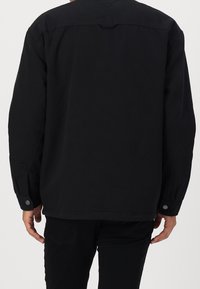 Svart jacka i mjukt tyg, med avslappnad passform, långa ärmar och silverfärgad knäppning vid ärmsluten. Enkelt, minimalistiskt design.