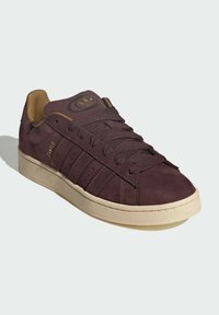 Maroon suede sneaker with beige accents, featuring textured laces, gold branding, and a rubber sole. Classic low-top design with stripes.