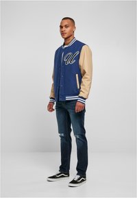 Jachetă de tip varsity albastră și bej cu broderie „U”, nasturi snap, manșete ribbate și guler plat, asociată cu blugi uzați și adidași negri.