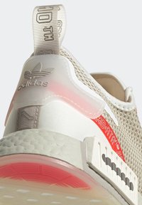 Ljust beige sneakers med ovandel i mesh, transparenta detaljer, röda accenter och strukturerad sula. Har en logotyp och en dragflik på hälen.