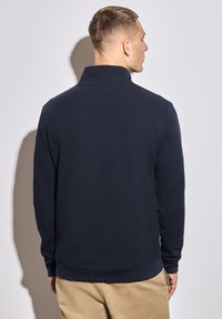 Muž otočený zády, oblečený v tmavě modrém svetru s dlouhým rukávem a sklopeným límcem, a béžových kalhotách, na jednoduchém světlém pozadí.