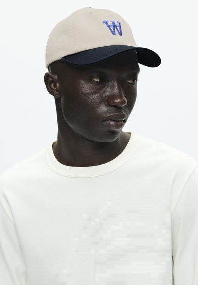 Casquette beige avec un pic marin, arborant un logo "W" bleu. Le tissu texturé complète une chemise à manches longues blanche à côtes.