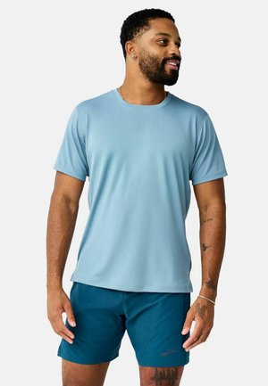Uomo con la barba che indossa una camicia azzurro chiaro a maniche corte e pantaloncini blu scuro, in piedi e guarda verso destra.
