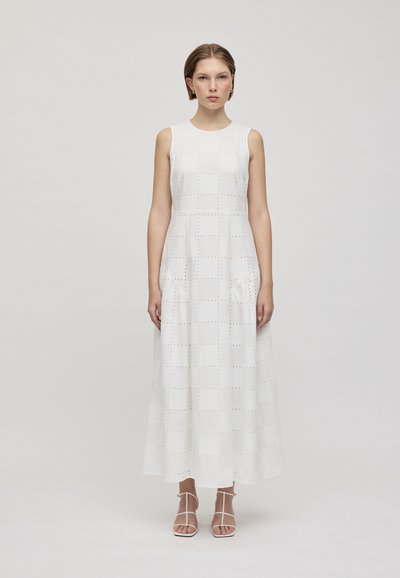 Robe maxi sans manches blanche avec un motif à carreaux et des découpes. Texture lisse, silhouette fluide et poches latérales. Portée avec des sandales à lanières.