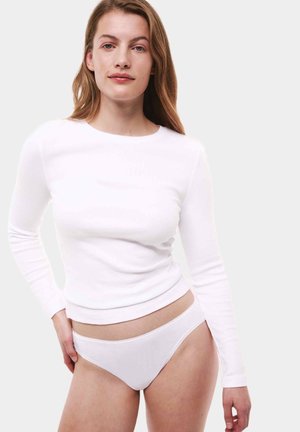 Vrouw in een witte lange mouwen shirt en witte ondergoed, staande met één hand op haar heup tegen een effen lichte achtergrond.