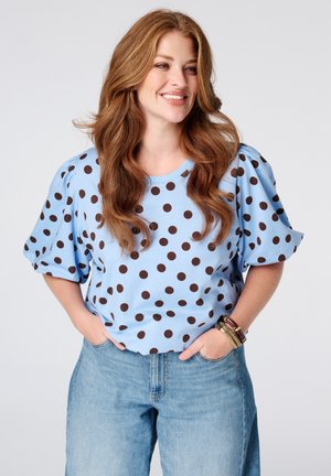 Femme souriante aux longs cheveux roux portant une blouse bleu clair à pois noirs et un jean bleu, les mains dans les poches, debout devant un fond blanc.