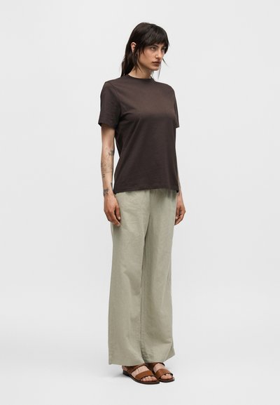 Γυναίκα με σκούρα μαλλιά που φοράει καφέ κοντομάνικο πουκάμισο, ανοιχτό μπεζ φαρδιά παντελόνια και καφέ σανδάλια, στέκεται μπροστά σε απλό φωτεινό φόντο.