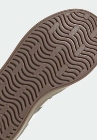 Brązowa gumowa podeszwa buta o falistej fakturze i okrągłych wzorach. Krawędź podeszwy lekko unosi się do góry, pokazując przejście do jasnej cholewki.