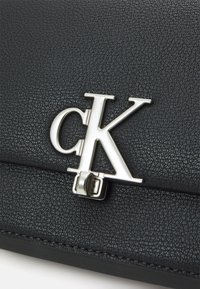 Borsa a mano in pelle nera con texture, caratterizzata da una chiusura con logo "ck" in argento e dettagli di cucitura visibili, che mette in mostra un design rettangolare strutturato.