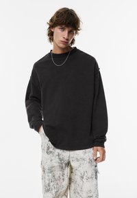 Zwarte oversized sweatshirt met lange mouwen, gestructureerde stof, ronde halslijn, gedragen met multikleurige cargo-broek met patroon. Zilveren ketting zichtbaar.