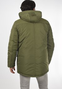 Parka acolchada de color verde olivo con capucha, hombros texturizados y puños ajustables, que presenta un dobladillo trasero más largo y bolsillos laterales.