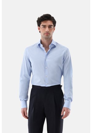 Uomo che indossa una camicia azzurro chiaro a maniche lunghe infilata in pantaloni neri eleganti, in piedi davanti a uno sfondo bianco neutro.