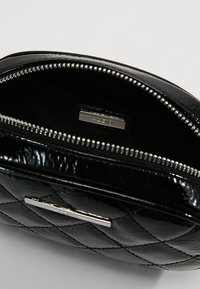 Borsa nera in pelle verniciata trapuntata con forma arrotondata. Presenta una zip argentata e una placca con logo metallico all'interno. Texture liscia, design compatto.