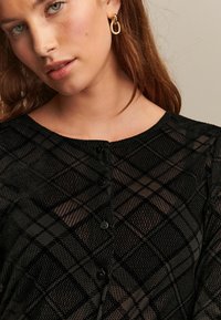 Černý cardigan se vzorem, který má kostkovaný design, kulatý výstřih, pět černých knoflíků a texturovanou tkaninu. Viditelné zlaté kruhové náušnice.