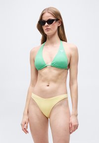 Γυναίκα που φοράει μαύρα γυαλιά ηλίου με σχήμα γάτας και μαγιό μπικίνι με πράσινο τοπ σε σχήμα τριγώνου και ανοιχτόκίτρινο κάτω μέρος, στέκεται μπροστά σε απλό φόντο.